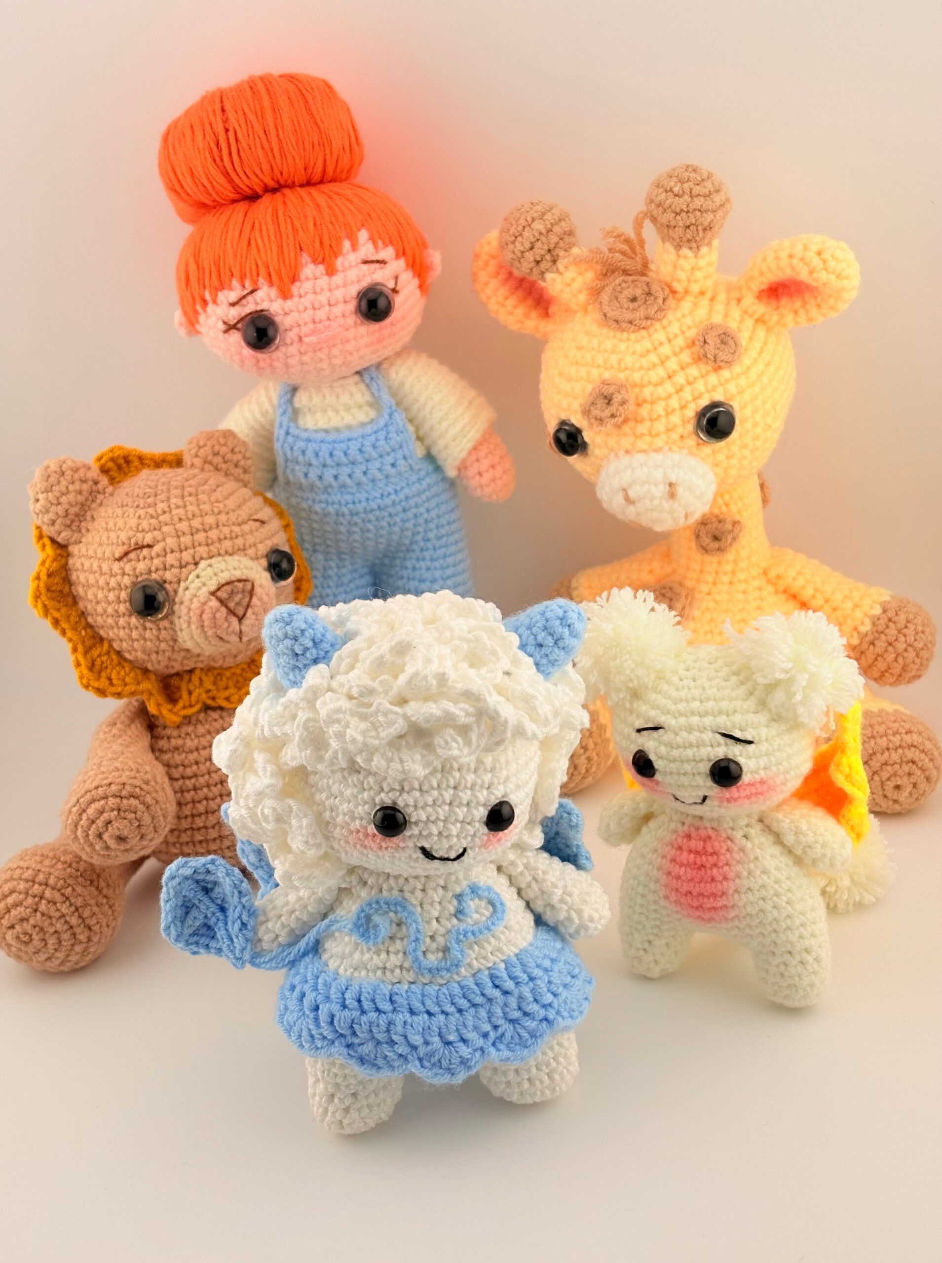 A group of five handmade crochet amigurumi dolls posed together against a light background. The figures include a girl with orange hair in a bun wearing blue overalls, a yellow giraffe with brown spots, a brown lion with a golden mane, a white and blue fluffy character with curly hair and horns, and a small cream-colored creature with orange and yellow wings. Each doll has black button eyes and a cute, cheerful expression.