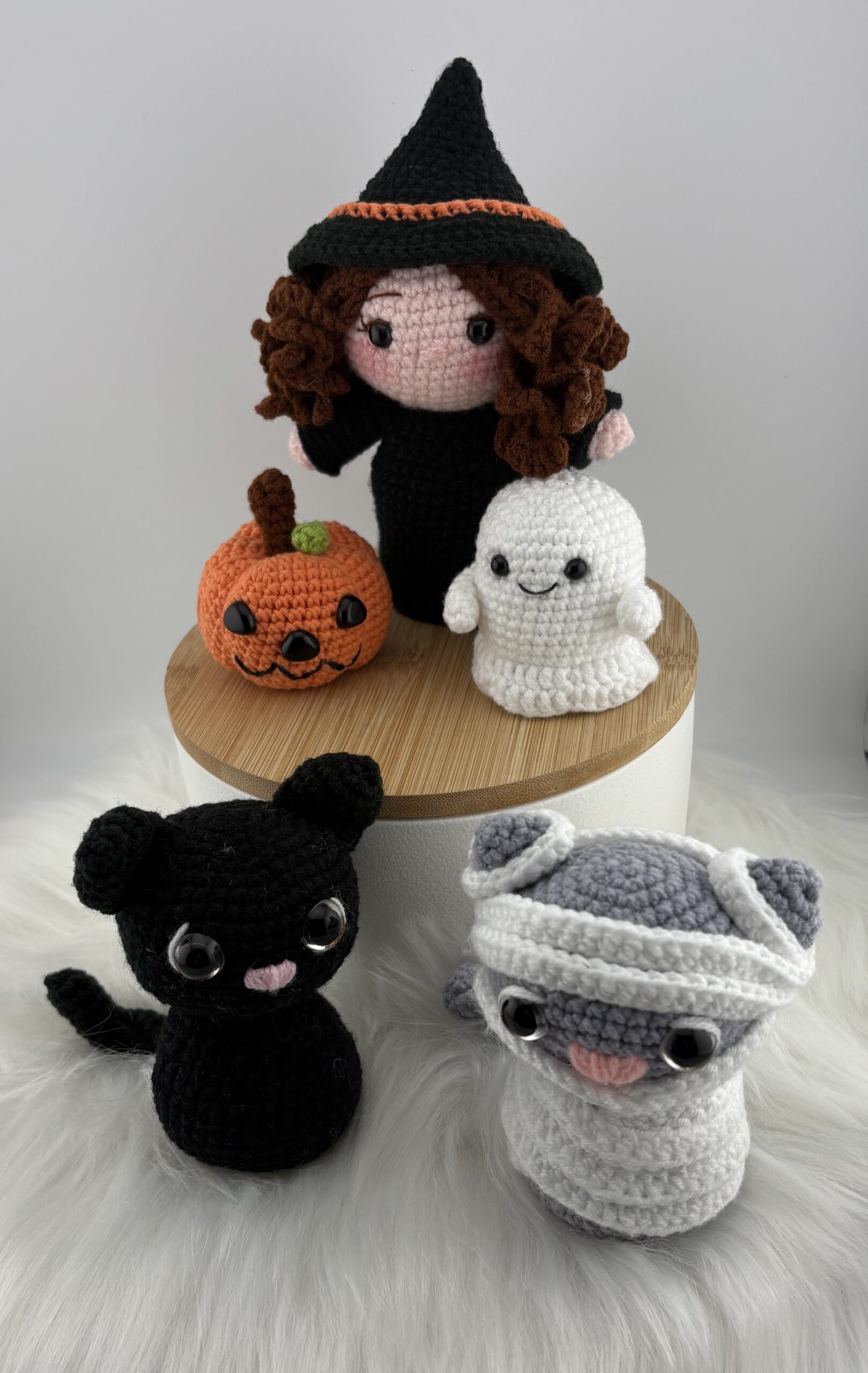 A handmade amigurumi Halloween collection featuring a witch doll with curly brown hair, a smiling pumpkin, a small white ghost, a black cat, and a gray mummy cat. All crocheted with soft yarn and detailed expressions.