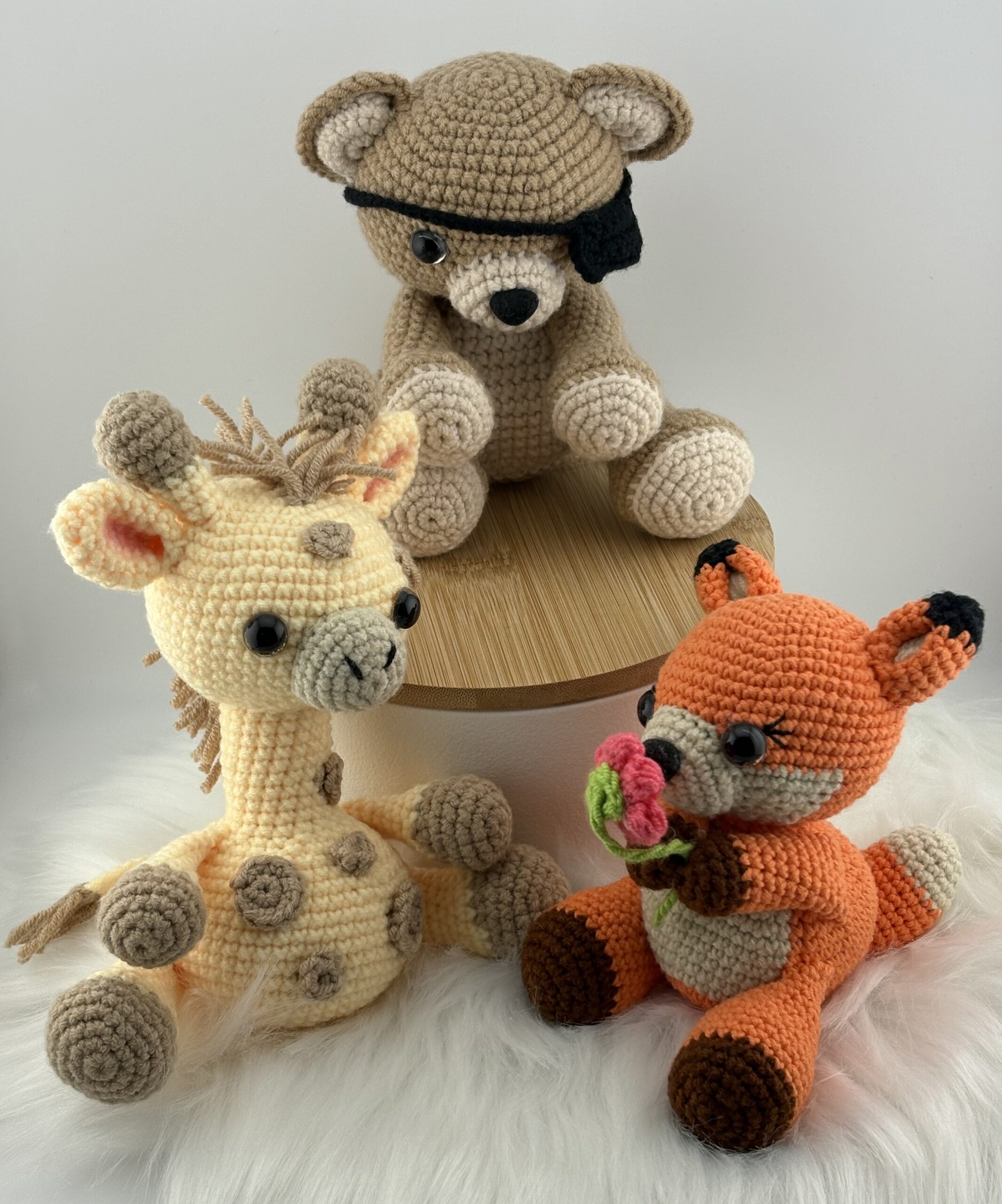 Three handmade amigurumi animals — a beige bear with an eye patch, a yellow giraffe, and an orange fox holding a pink flower — displayed on a wooden stand and white fur background.