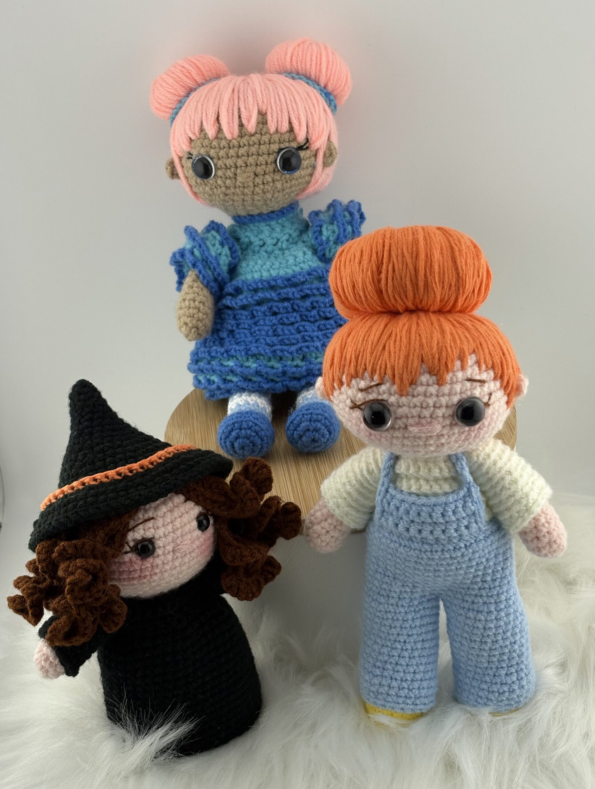 Three crocheted amigurumi dolls displayed together: one with pink hair in a blue dress, one with orange hair in overalls, and a witch doll in black with curly brown hair and a pointed hat. The dolls are handmade with yarn and safety eyes.