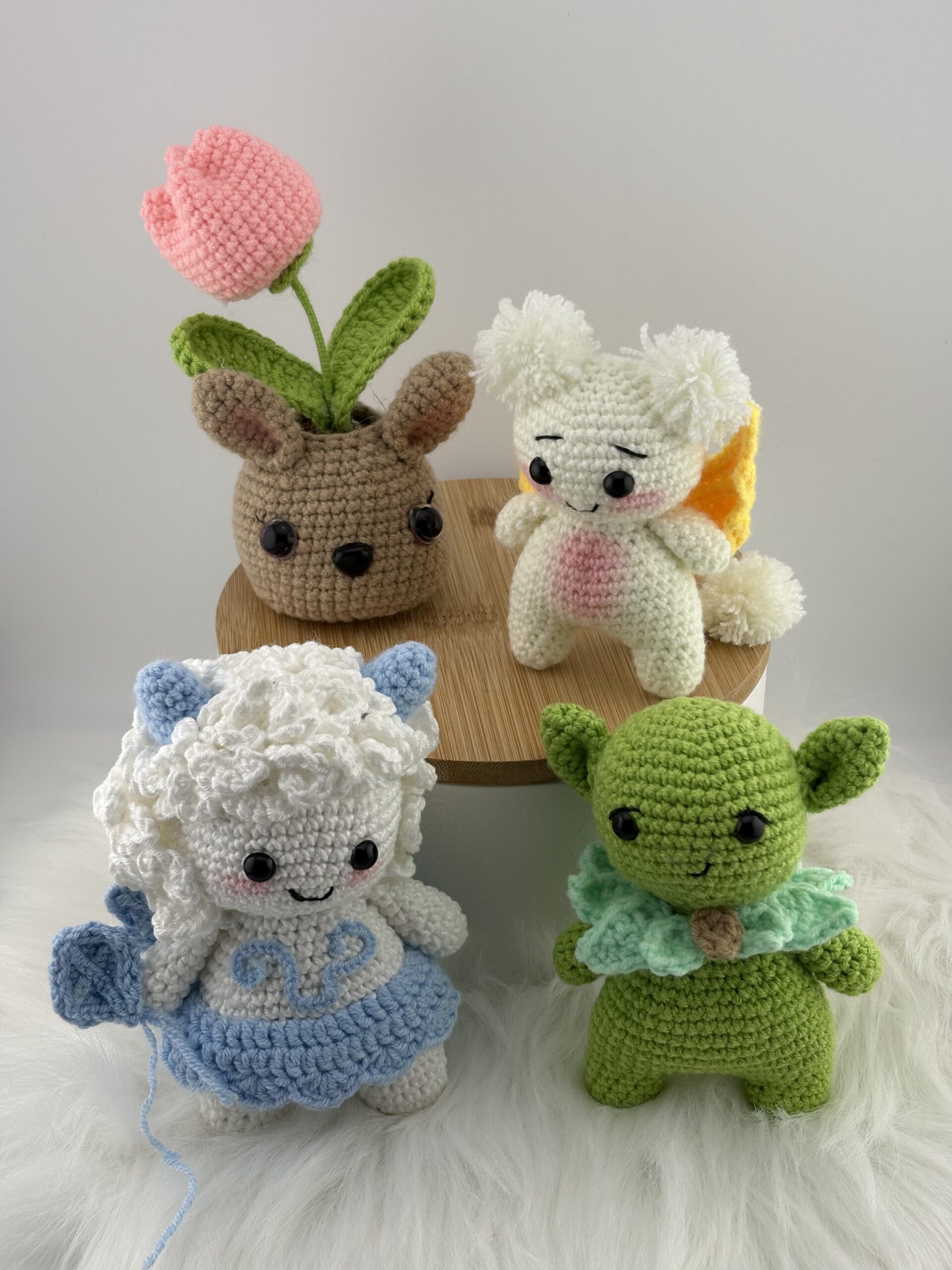 Four cute crocheted amigurumi characters — a bunny flower pot with a pink tulip, a white creature with wings, a blue curly-haired lamb, and a small green elf — arranged on a wooden stand over a fluffy white surface.