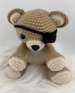 A handmade crochet teddy bear amigurumi with light brown and beige yarn. The bear is sitting upright and wearing a small black eye patch over one eye, giving it a playful pirate look. It has black safety eyes, a black embroidered nose, and a soft, cozy appearance against a plain white background.