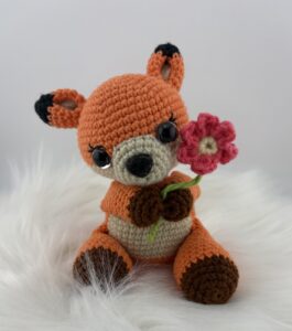 A crocheted orange fox amigurumi with big black eyes and brown paws is holding a pink and white crocheted flower. The fox has black-tipped ears and a beige snout and belly, sitting upright against a plain white background.
